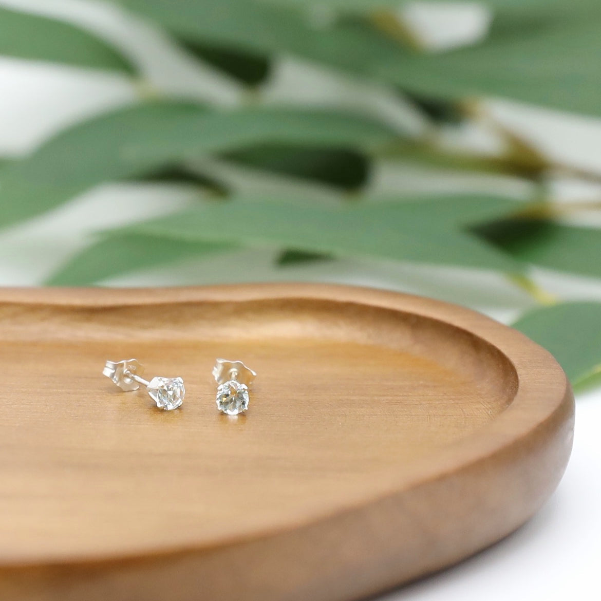 A pair of Light Green Emerald stud earrings placed on a woden tray with eucalyptus leaves on the background.