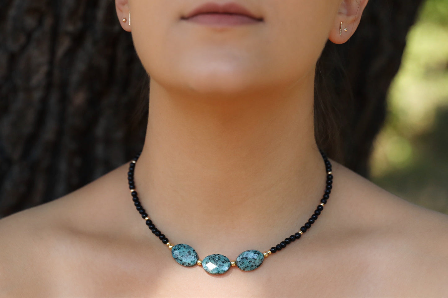 A model wearing The Fortifying Foundations Necklace by Gaiai's Heirlooms, made with Black Onyx beads, three Blue Spot Jasper stones in the middle and Gold Filled details.