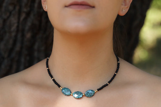 A model wearing The Fortifying Foundations Necklace by Gaiai's Heirlooms, made with Black Onyx beads, three Blue Spot Jasper stones in the middle and Gold Filled details.